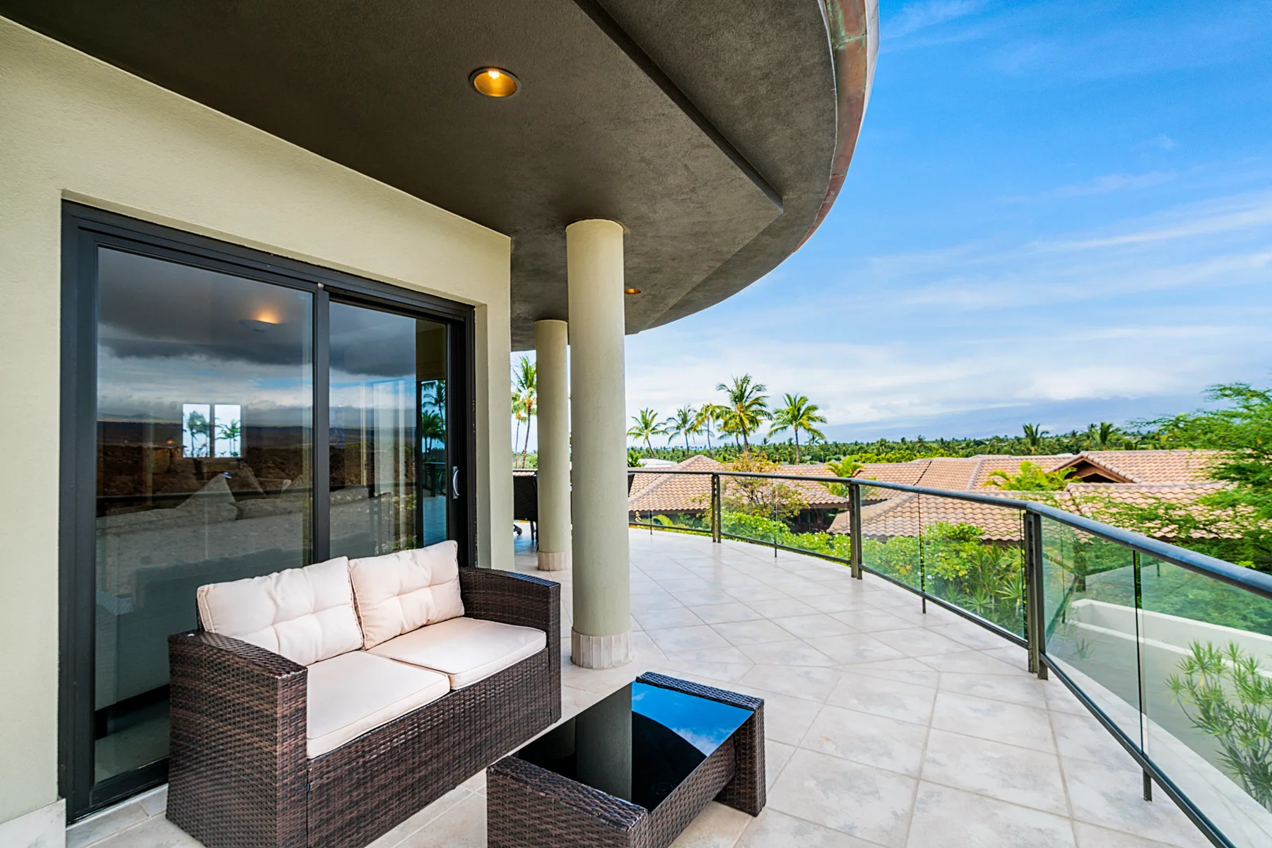 A spacious tiled balcony with outdoor seating and glass railings overlooks palm trees and rooftops.