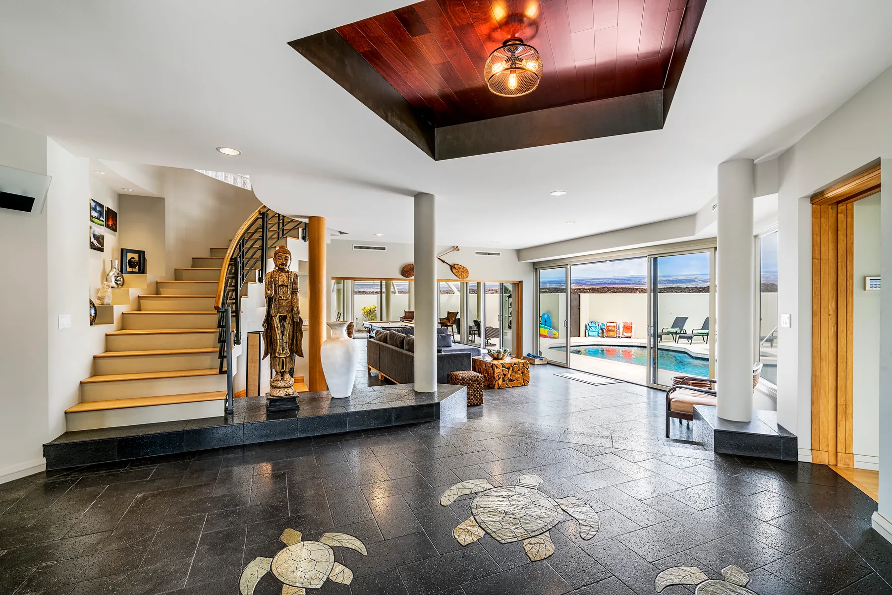 A bright living area with a staircase, dark tiled floor, and large windows overlooking a pool.