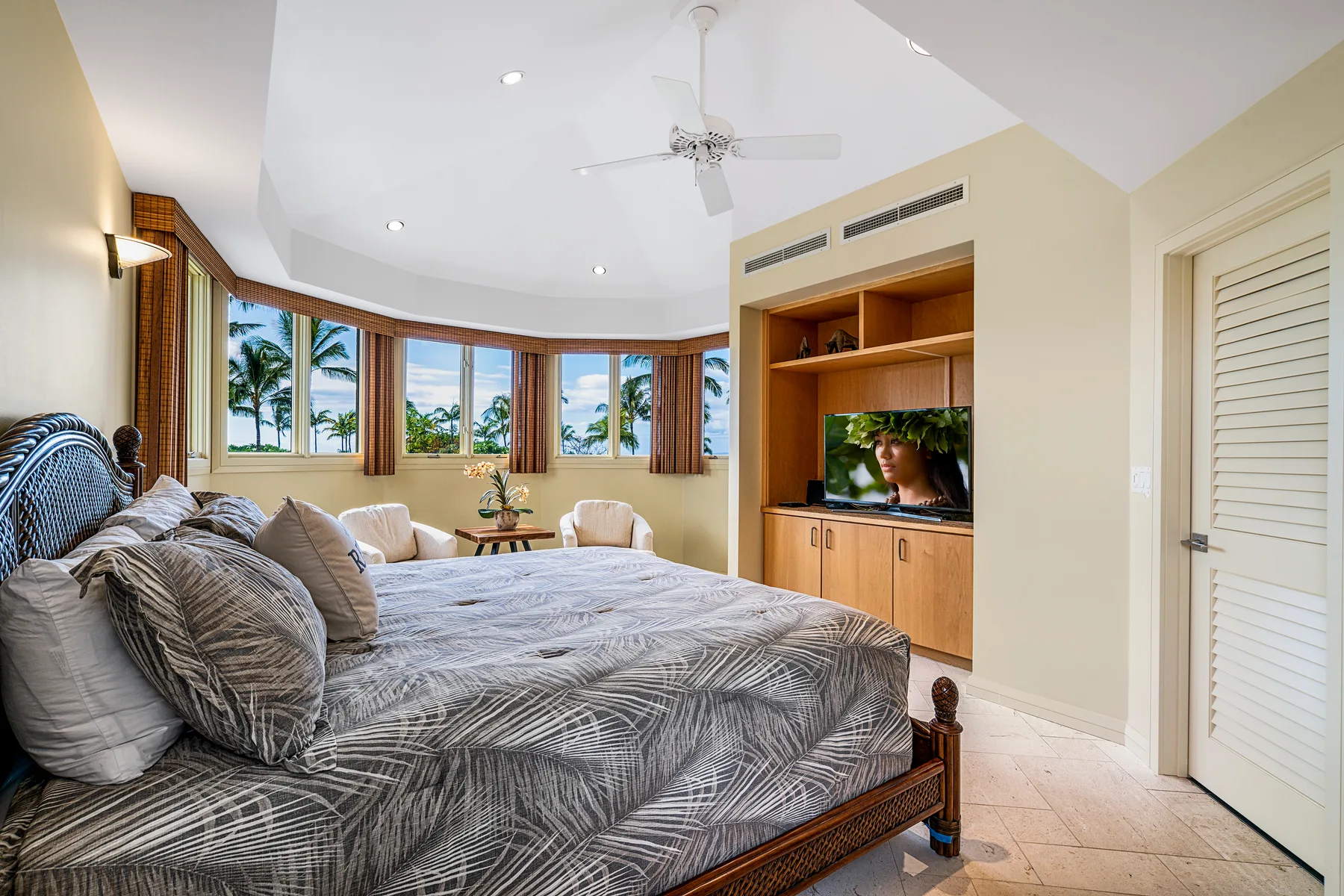 A bedroom with a large bed, a television, and windows overlooking palm trees.