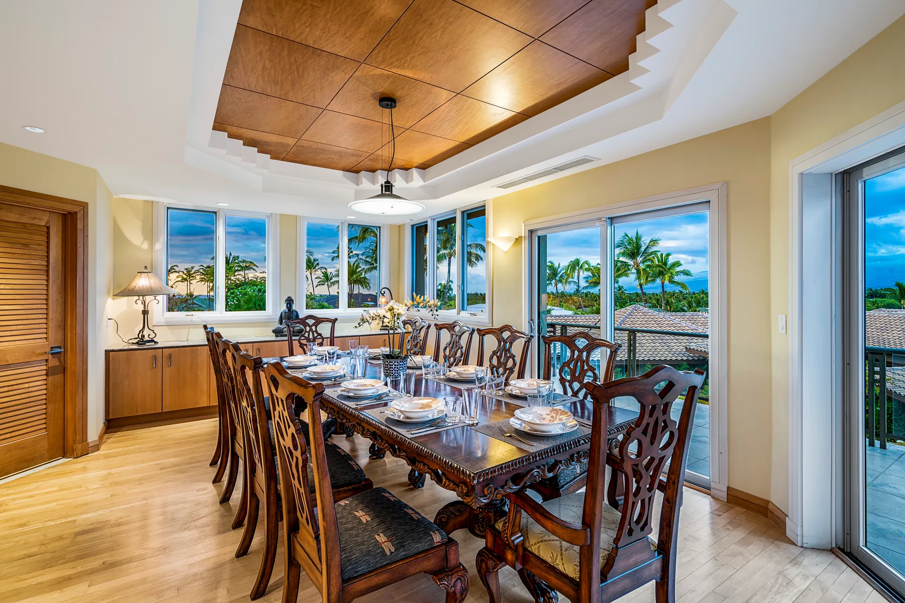 A dining room with a long table set beside windows overlooking palm trees and water.