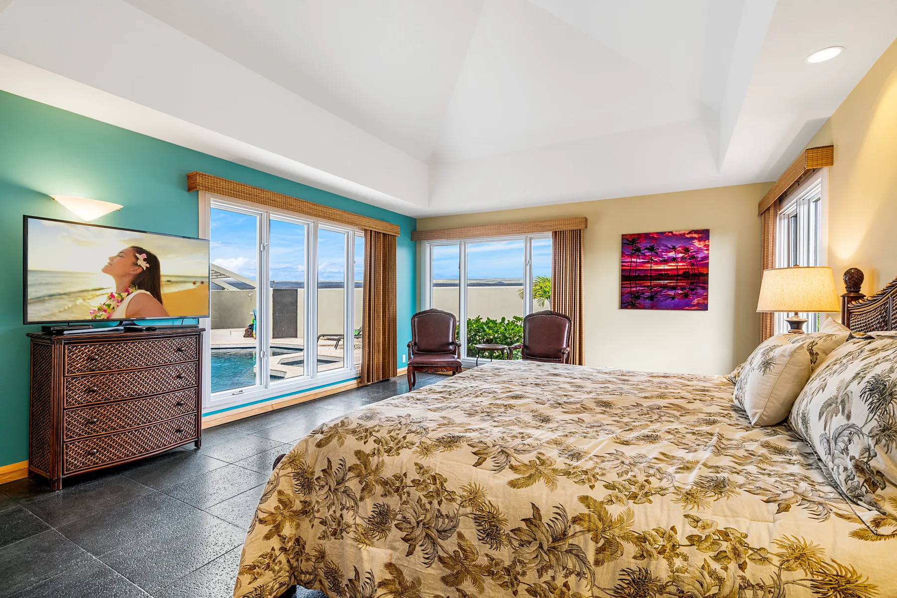 A bright bedroom has a large bed with a floral bedspread and windows overlooking the water.