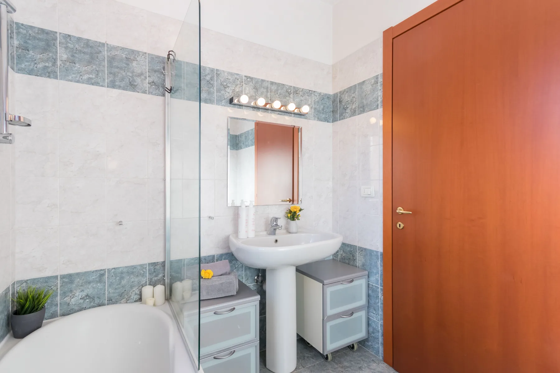 Un baño alicatado con bañera, mampara de ducha de vidrio, lavabo y puerta de madera.