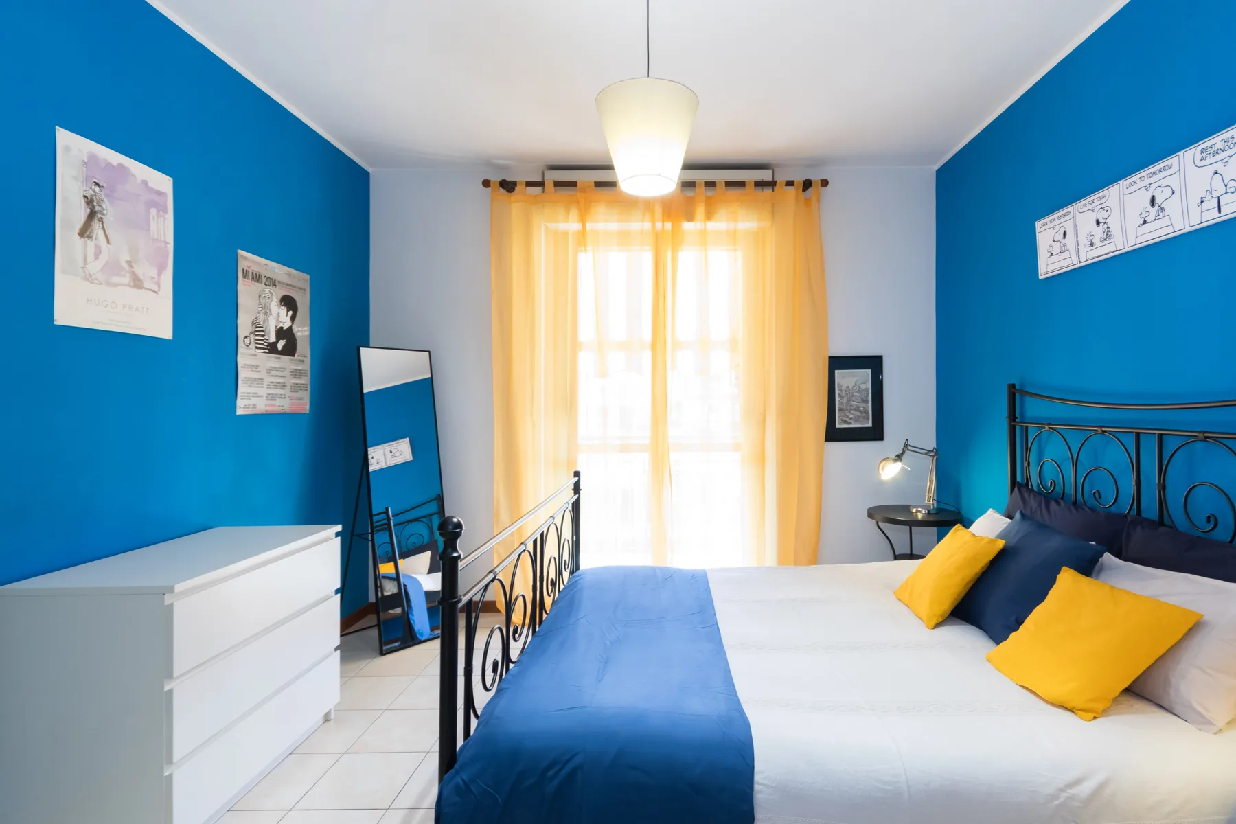 Un dormitorio con una cama, paredes azules y una ventana luminosa con cortinas amarillas.