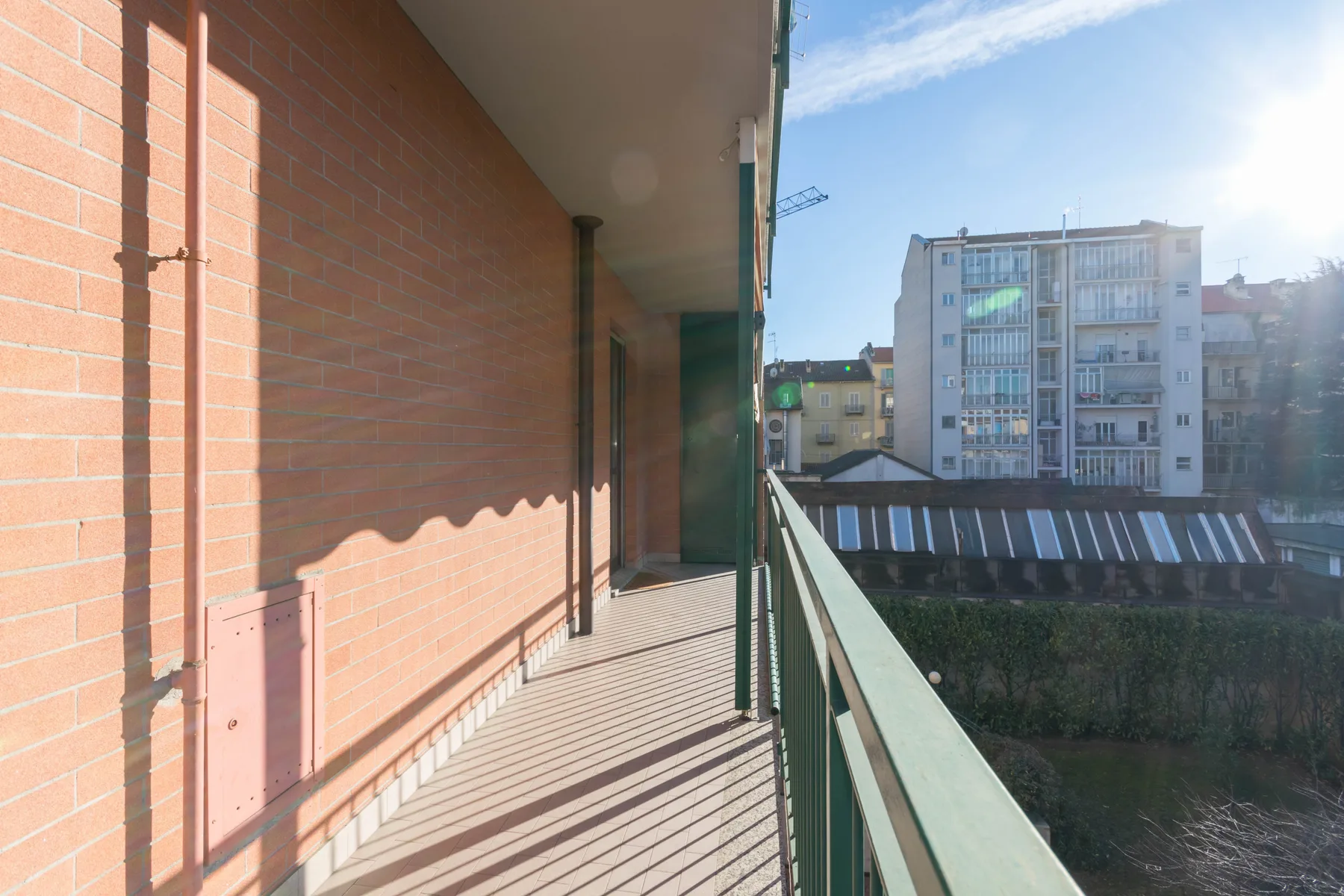 Un balcón estrecho con pared de ladrillo y barandilla verde da a edificios cercanos.