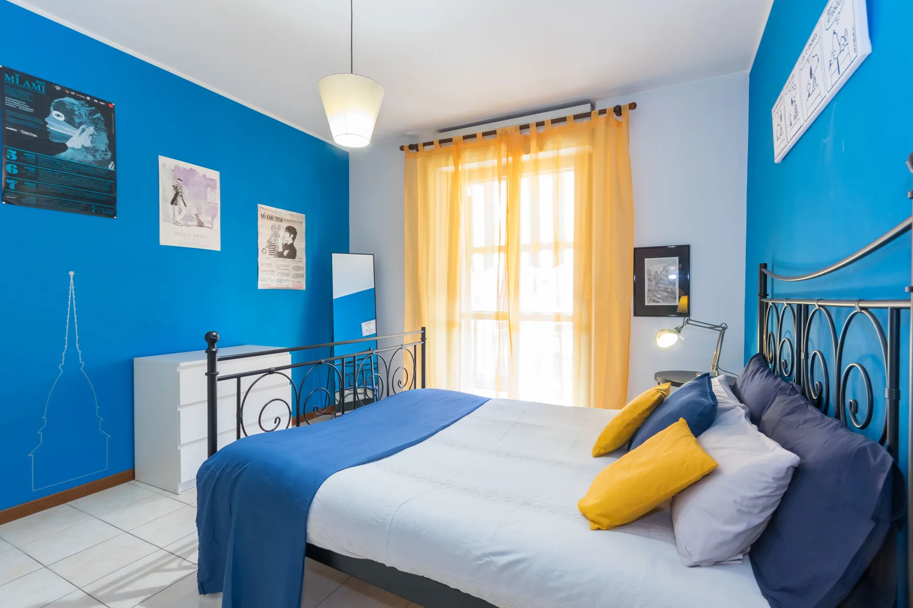 Un dormitorio luminoso con paredes azules, una cama y luz que entra por cortinas amarillas.