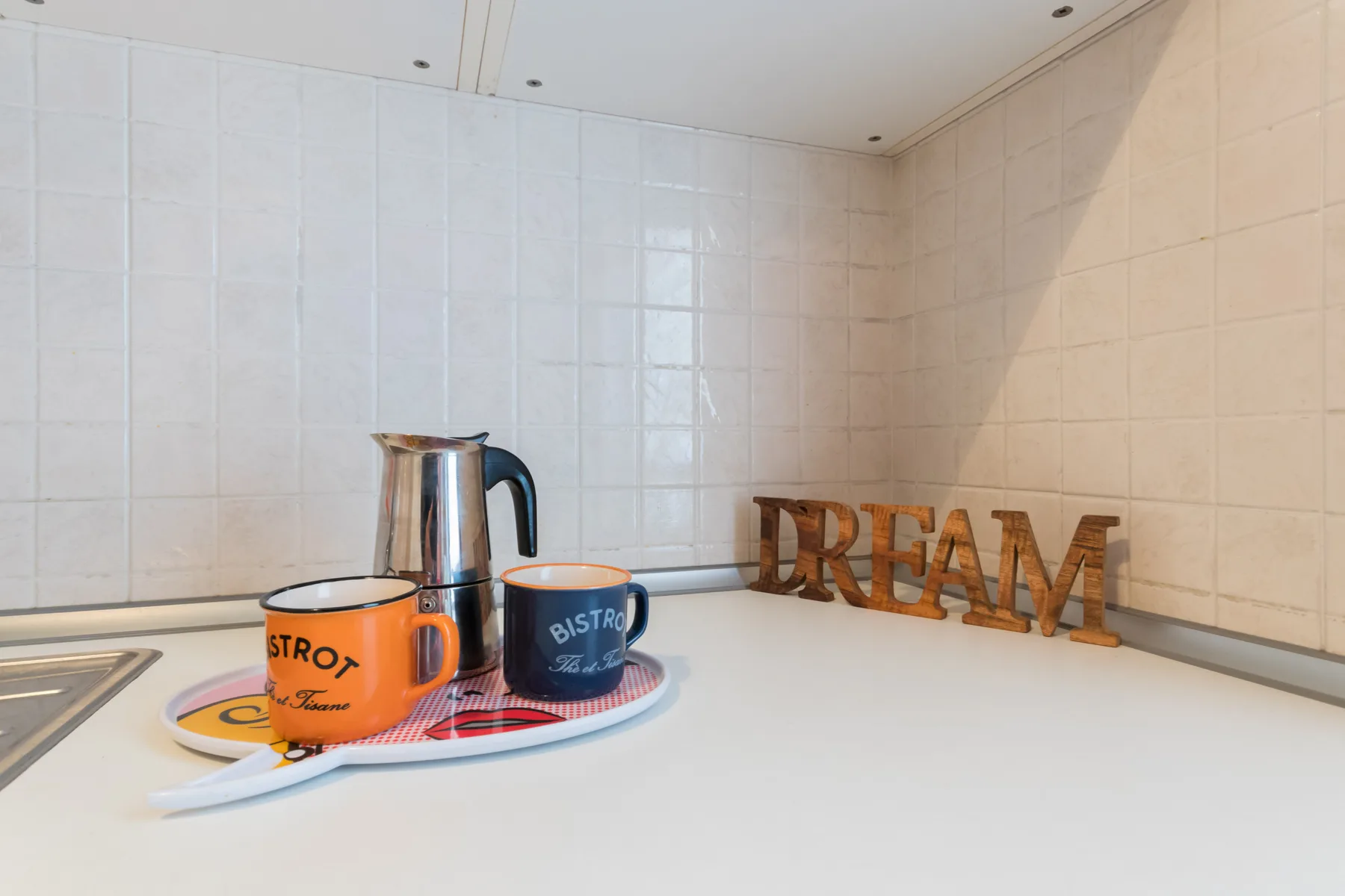 Una encimera de cocina alicatada sostiene dos tazas, una tetera pequeña y letras de madera.