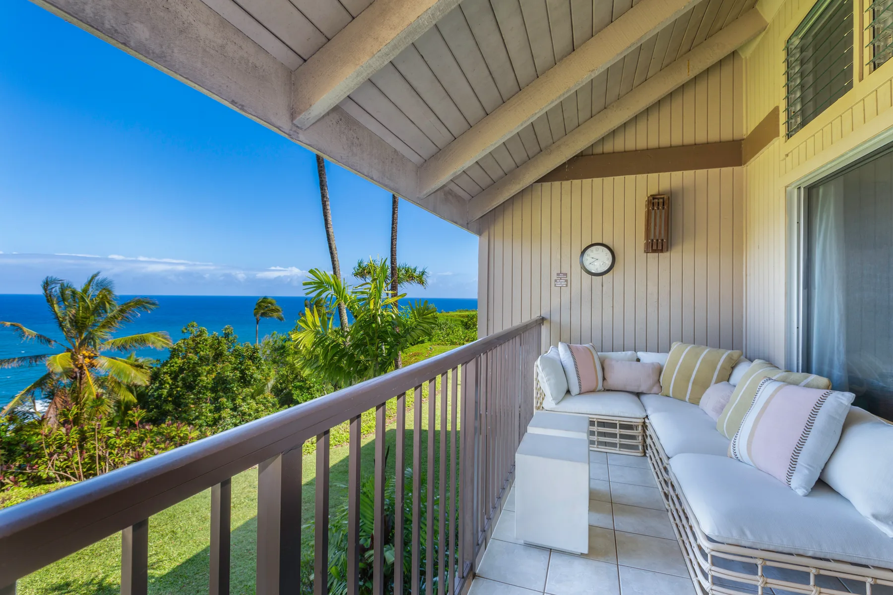A covered balcony with cushioned seating overlooks palm trees and the ocean.