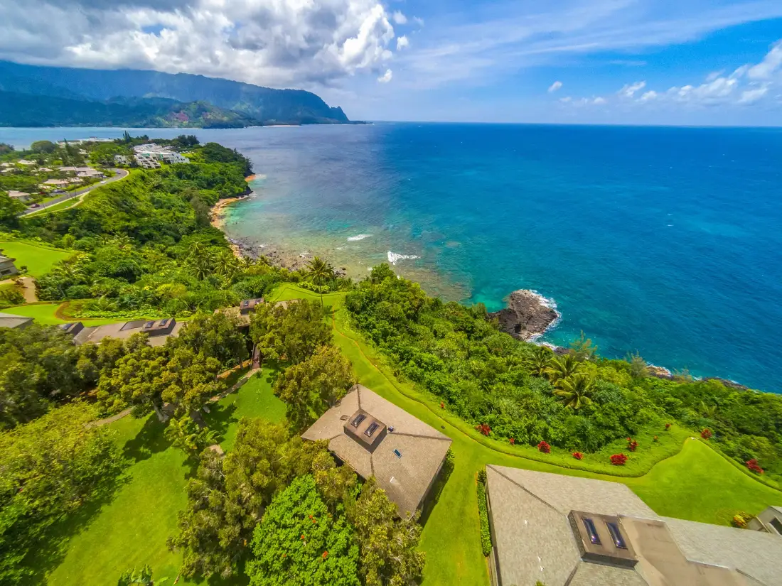 Aerial view of a green coastline beside blue ocean water and distant mountains.