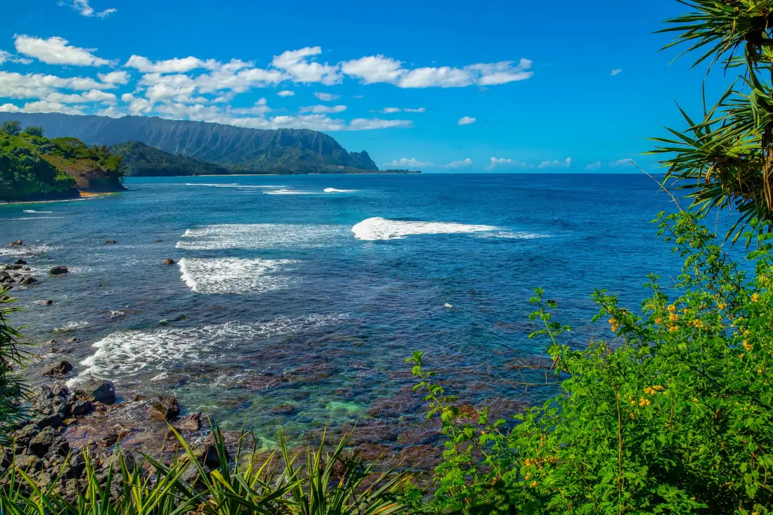 A coastal seascape with waves breaking over a shallow reef beneath a blue sky.