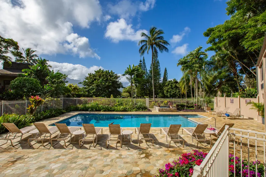 A swimming pool is surrounded by lounge seating, tropical greenery, and a paved patio.