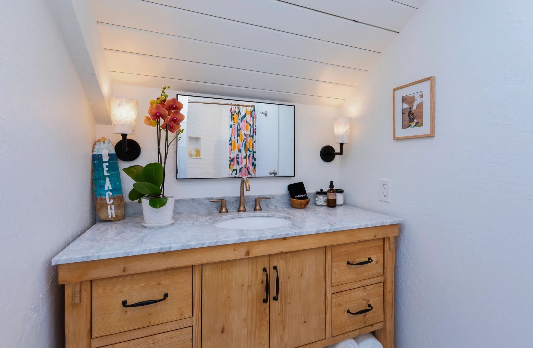 A wooden bathroom vanity sits beneath a mirror with colorful artwork.