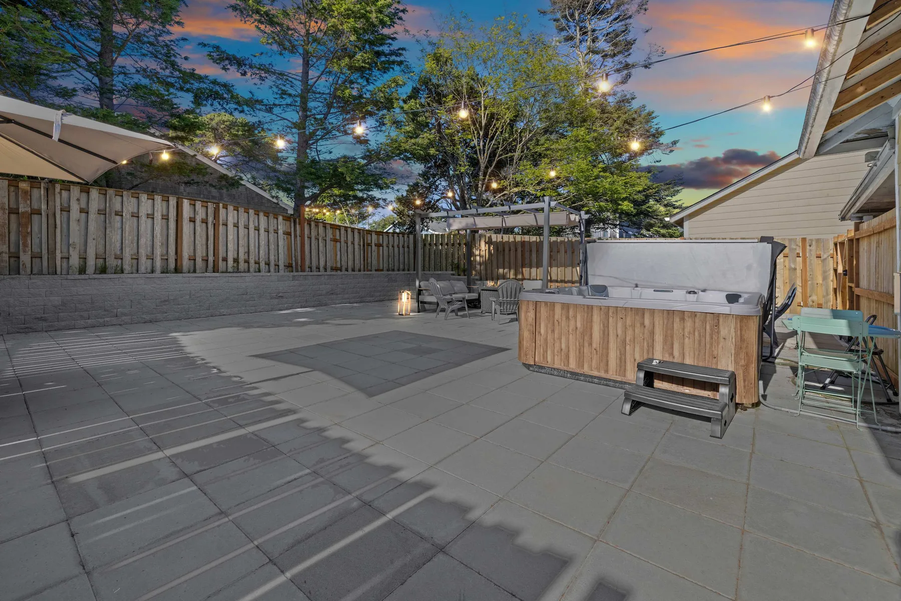 A paved outdoor patio with a hot tub and seating beneath string lights.