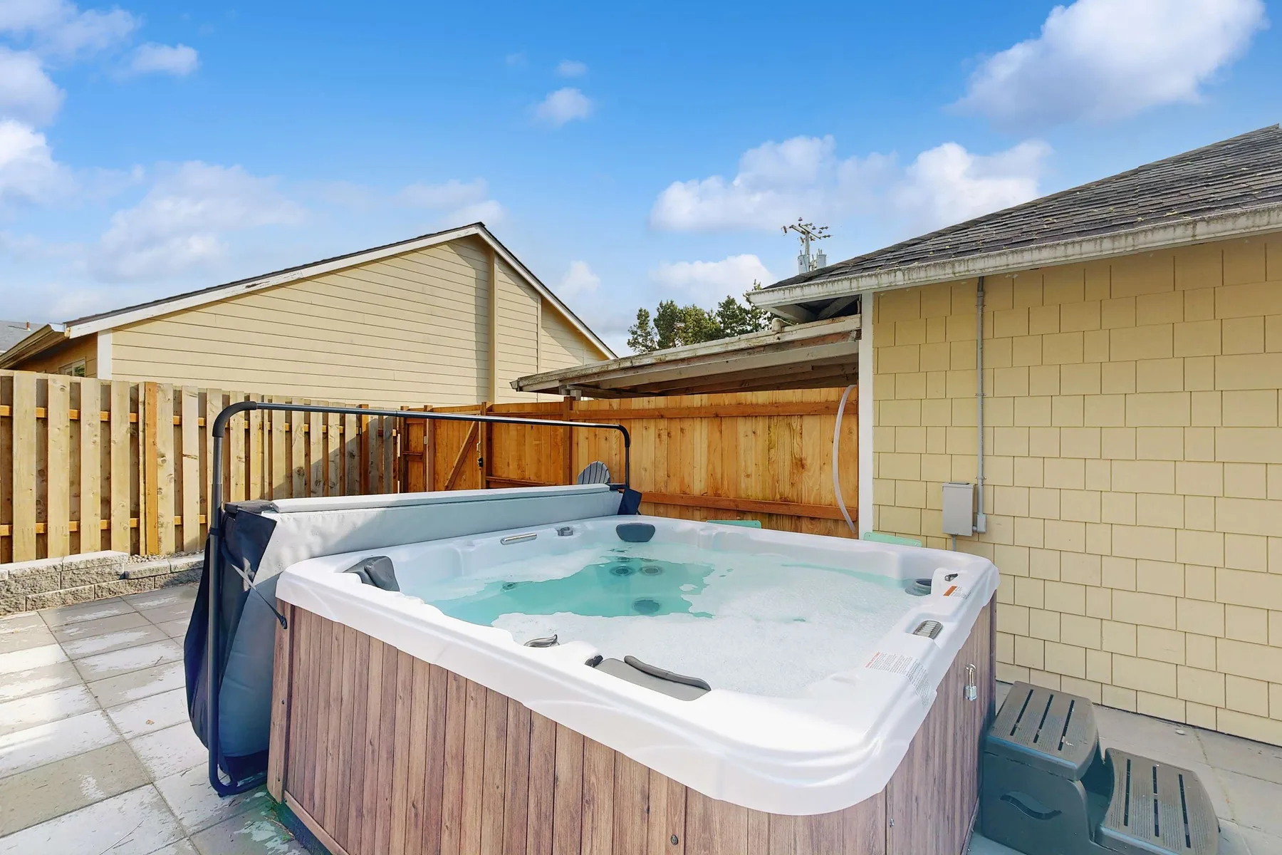 An outdoor hot tub sits beside a fenced patio under a blue sky.