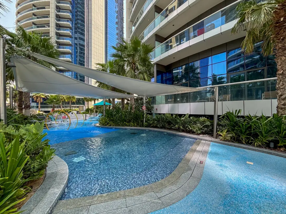 Una piscina exterior ajardinada está bajo toldos de sombra junto a edificios modernos.