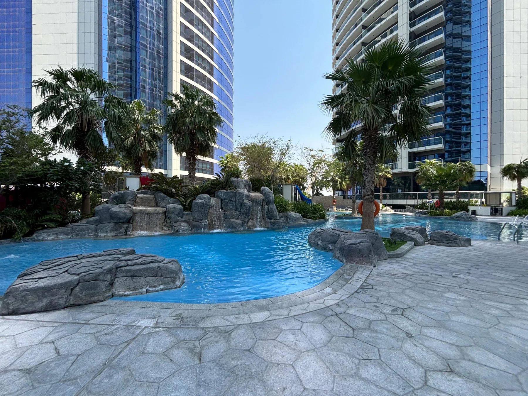 Una piscina al aire libre con rocas está entre edificios altos y palmeras.