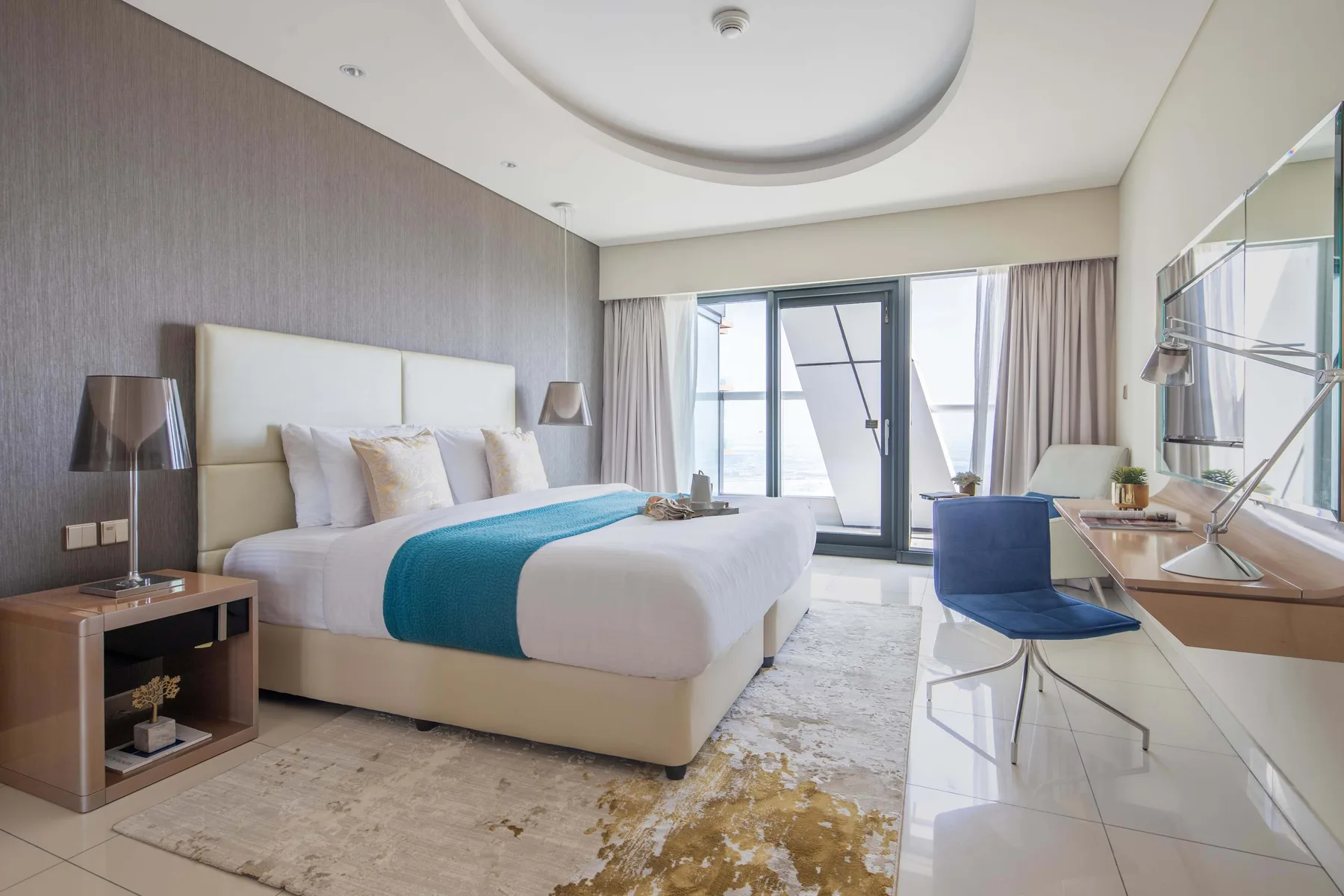 Un dormitorio con una cama bien hecha, una manta azul y grandes ventanas.
