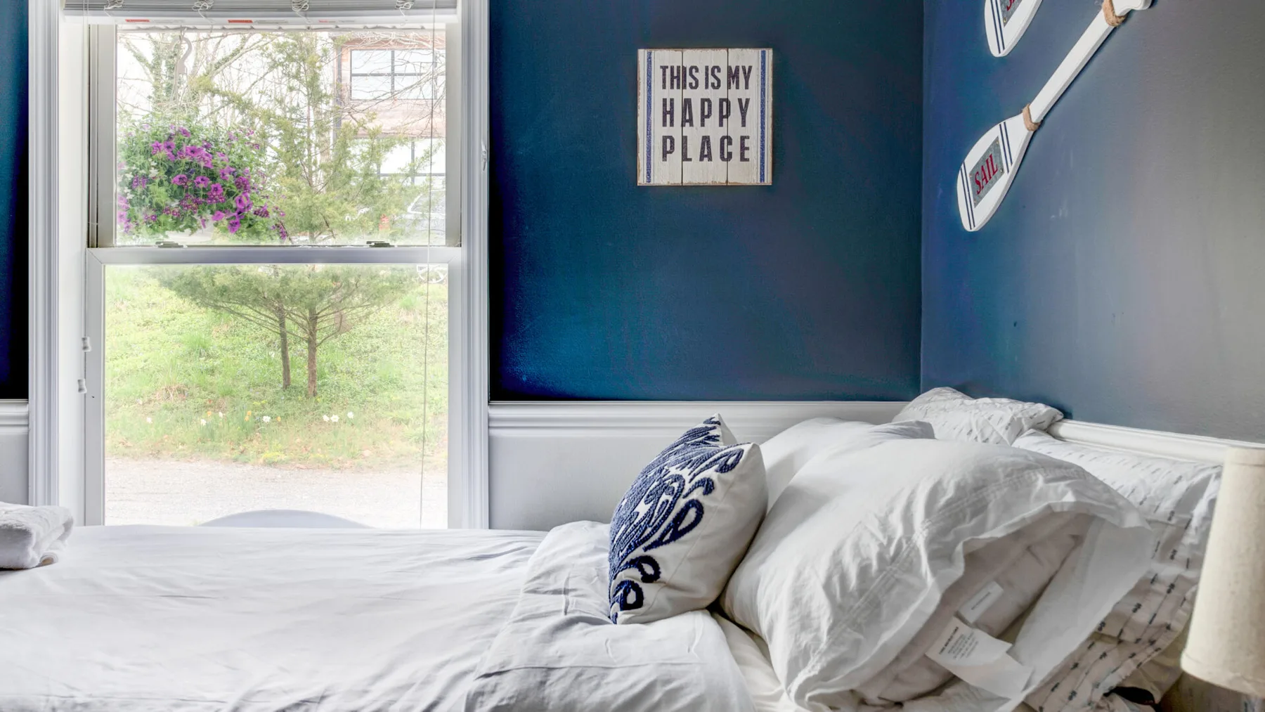 A bedroom with white bedding, blue walls, and a large window overlooking greenery.
