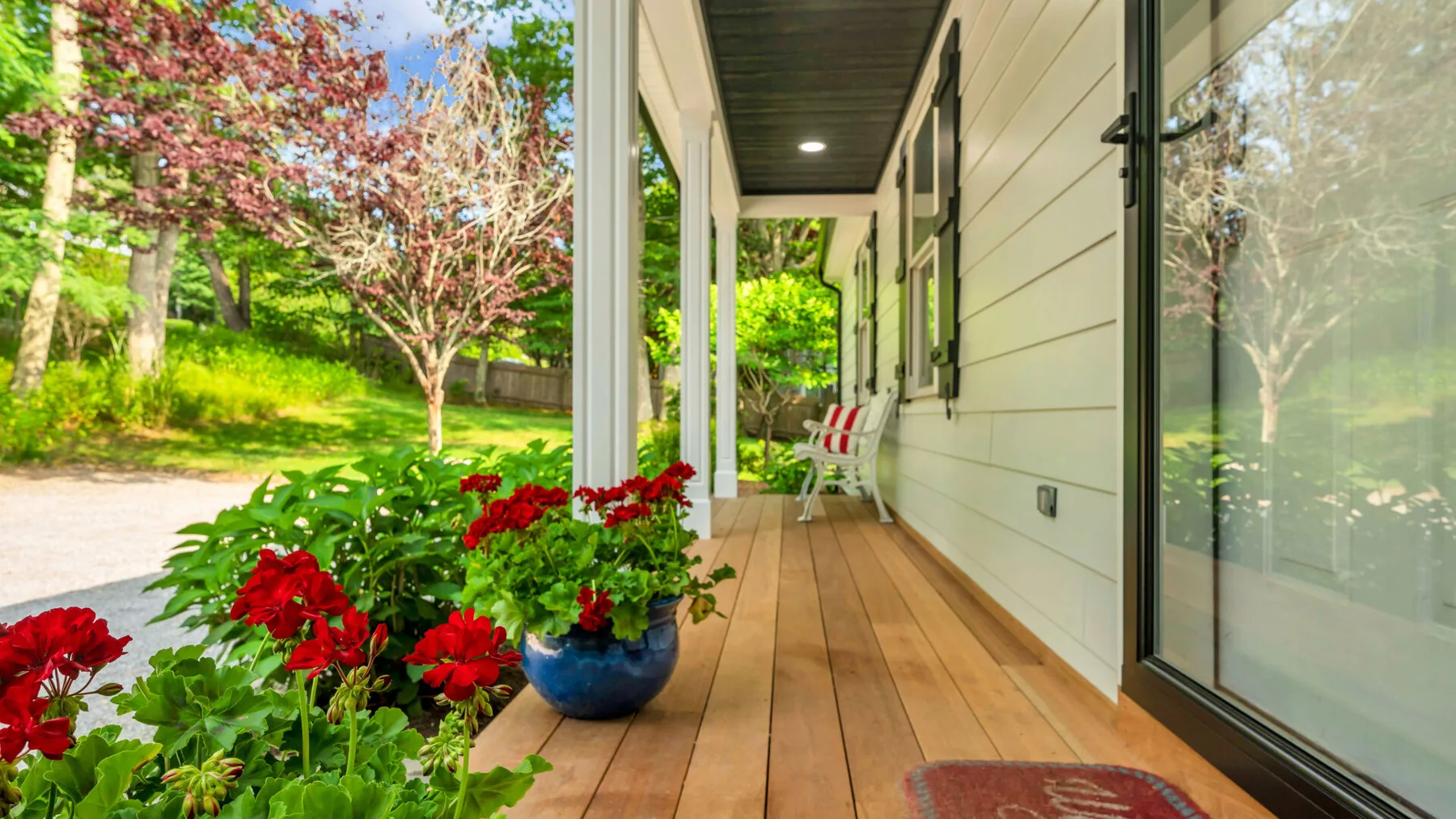 A covered porch with a wooden deck, glass door, and potted flowers overlooks a leafy yard.
