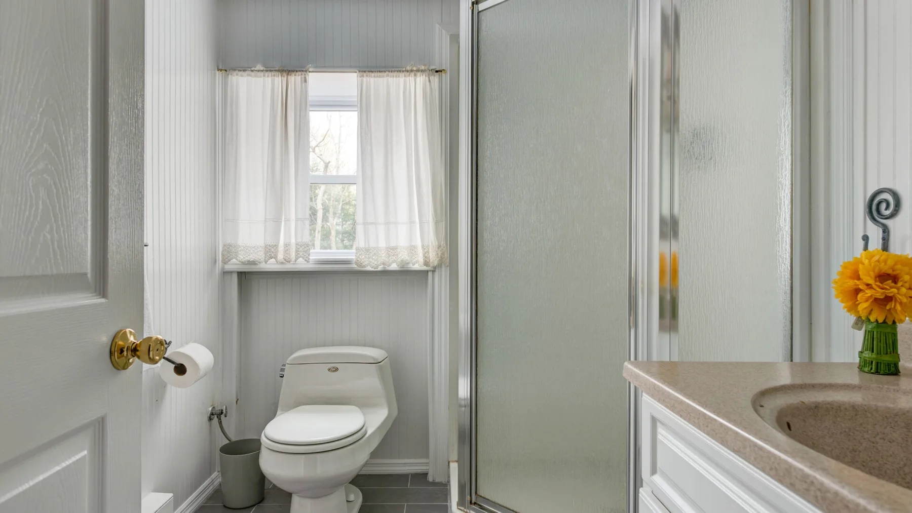A bathroom with a toilet, shower, and sink beneath a curtained window.