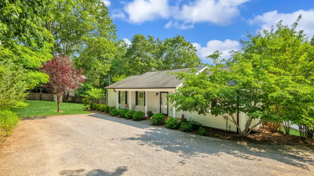 A low house stands beside a broad gravel driveway, surrounded by leafy trees.