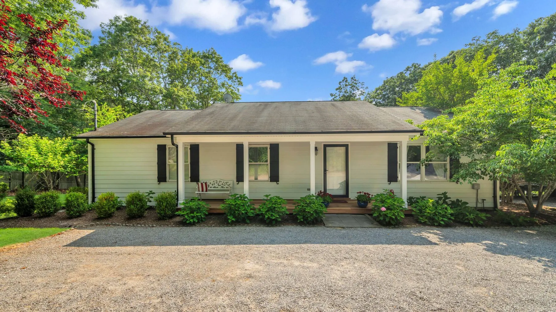 A single-story house stands behind a broad gravel driveway, surrounded by trees and shrubs.