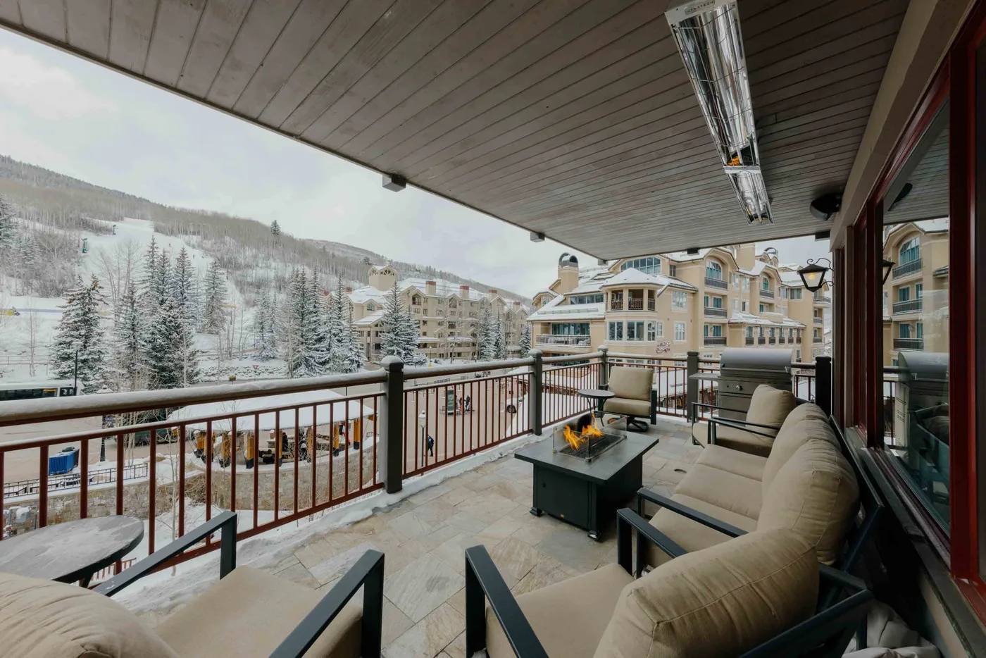 A covered outdoor patio with seating overlooks snow-covered trees and nearby buildings.