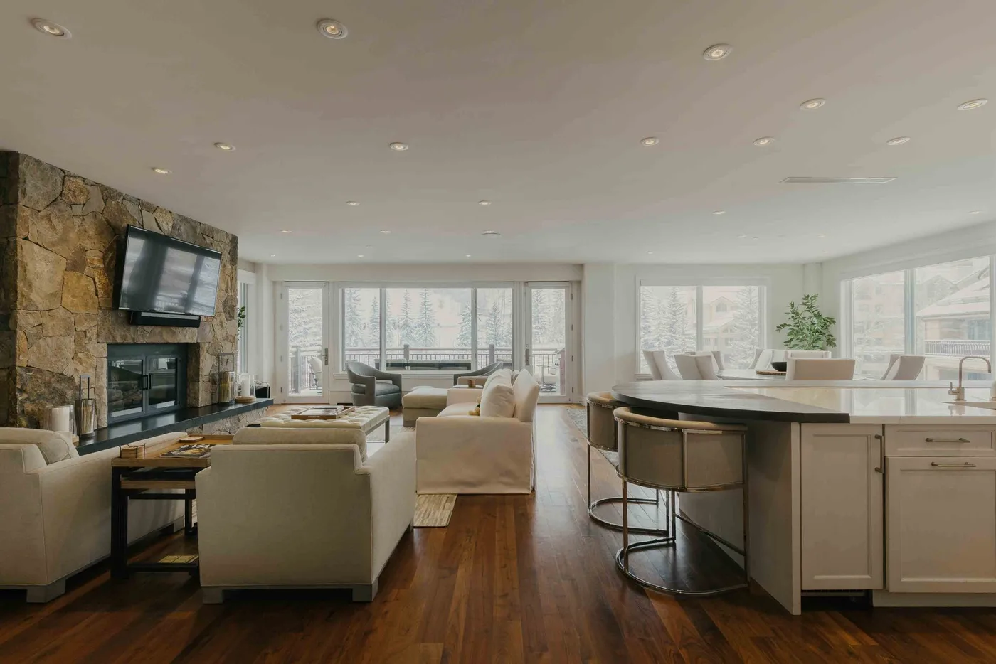 A bright open-plan living room and kitchen with large windows and a stone fireplace.