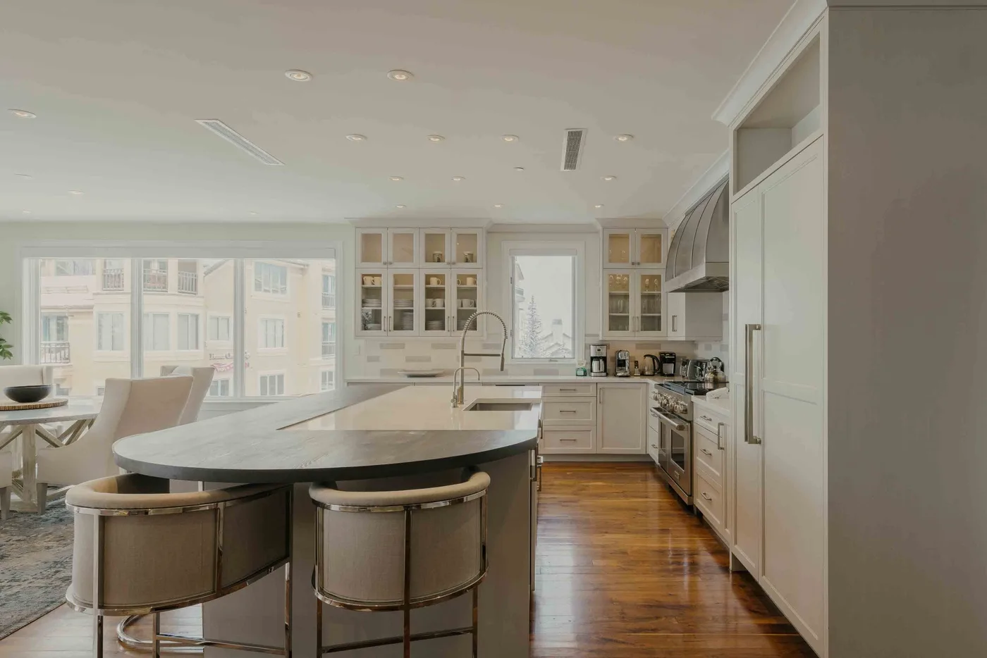 A bright kitchen with a large island, white cabinetry, and windows overlooking nearby buildings.