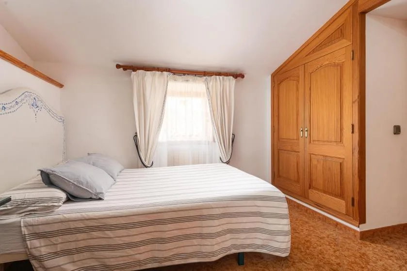 A bedroom with a striped bed, a curtained window, and a wooden wardrobe.