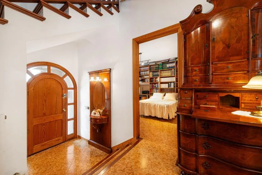 A warm-toned interior hallway leads past wooden furnishings to a bedroom with bookshelves.