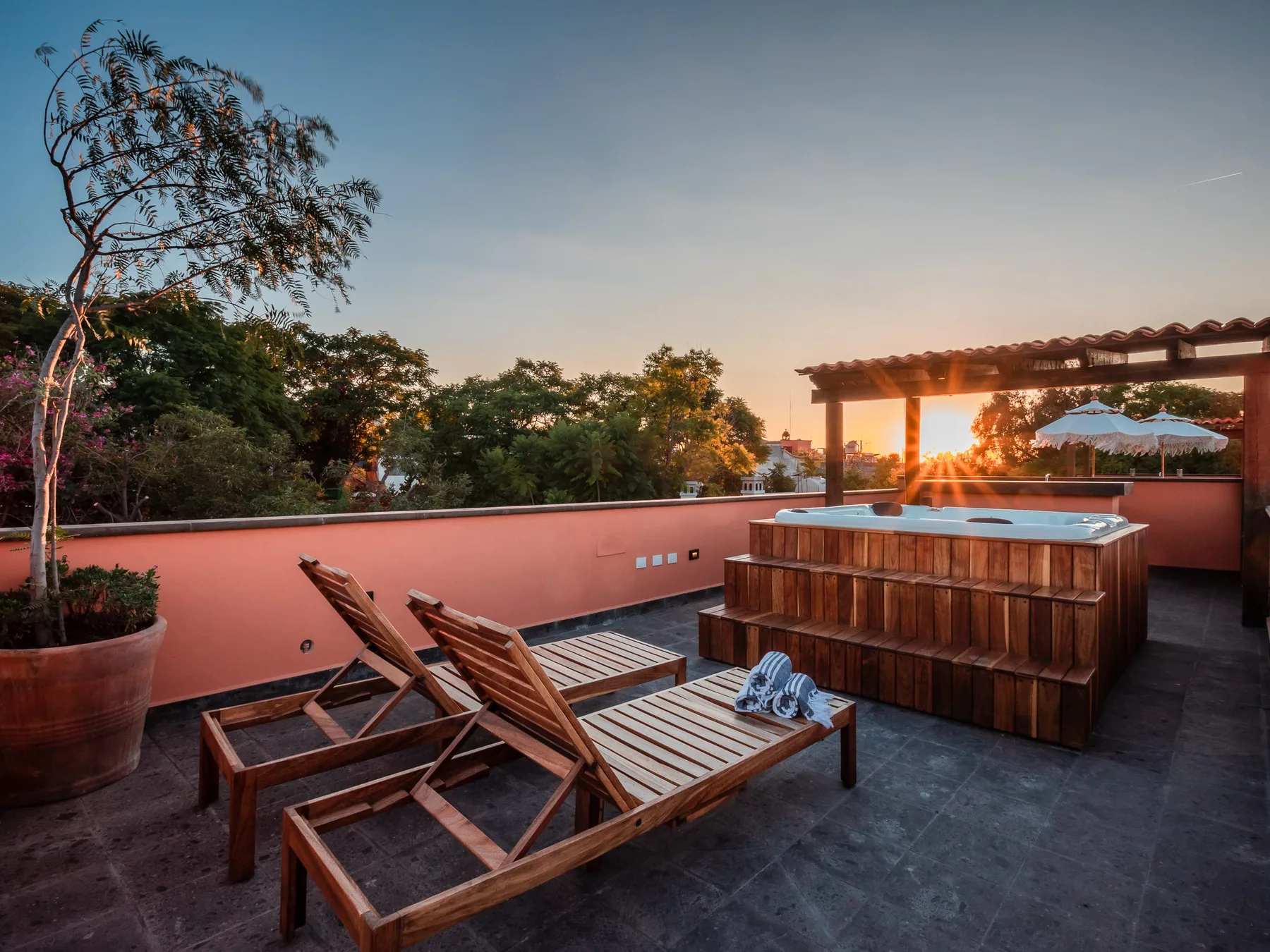 A rooftop terrace with a wooden hot tub and reclining lounge chairs at sunset.