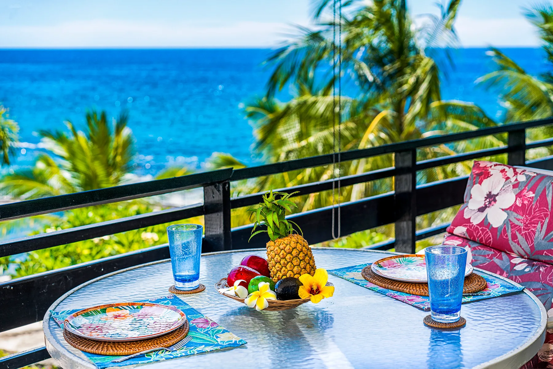 Una mesa de comedor en un balcón mira al océano, con fruta tropical y vajilla colorida.