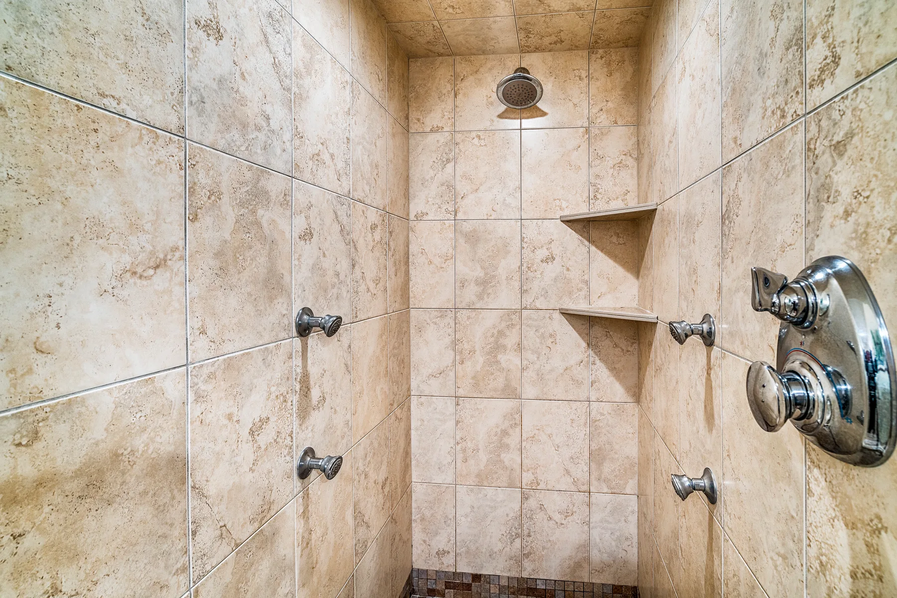 Una ducha con azulejos, un cabezal montado en la pared y estantes en las esquinas.