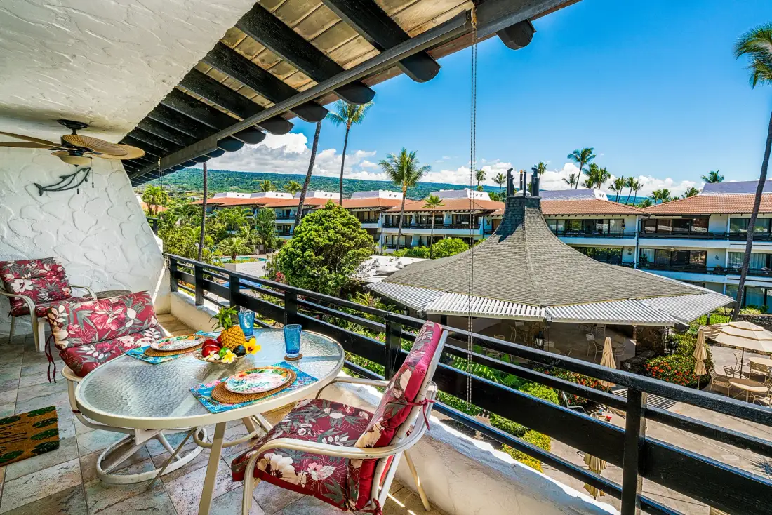 Un balcón cubierto tiene una mesa pequeña y asientos con vistas a un patio tropical.