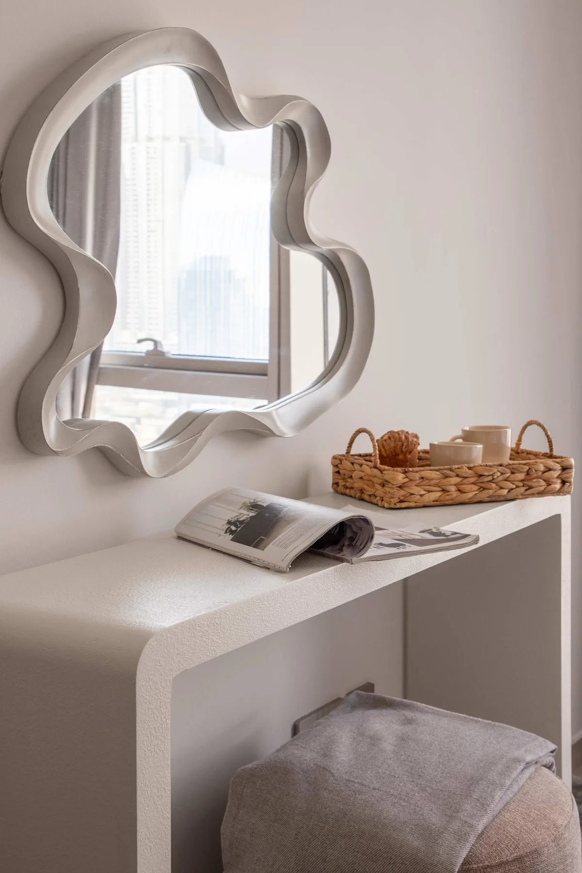 Un tocador de dormitorio con un espejo decorativo y una bandeja tejida sobre la superficie.