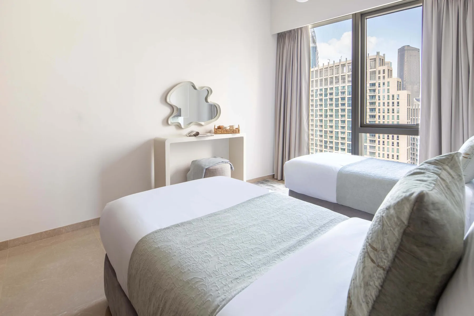 Un dormitorio luminoso con dos camas junto a una ventana grande con vistas a edificios.