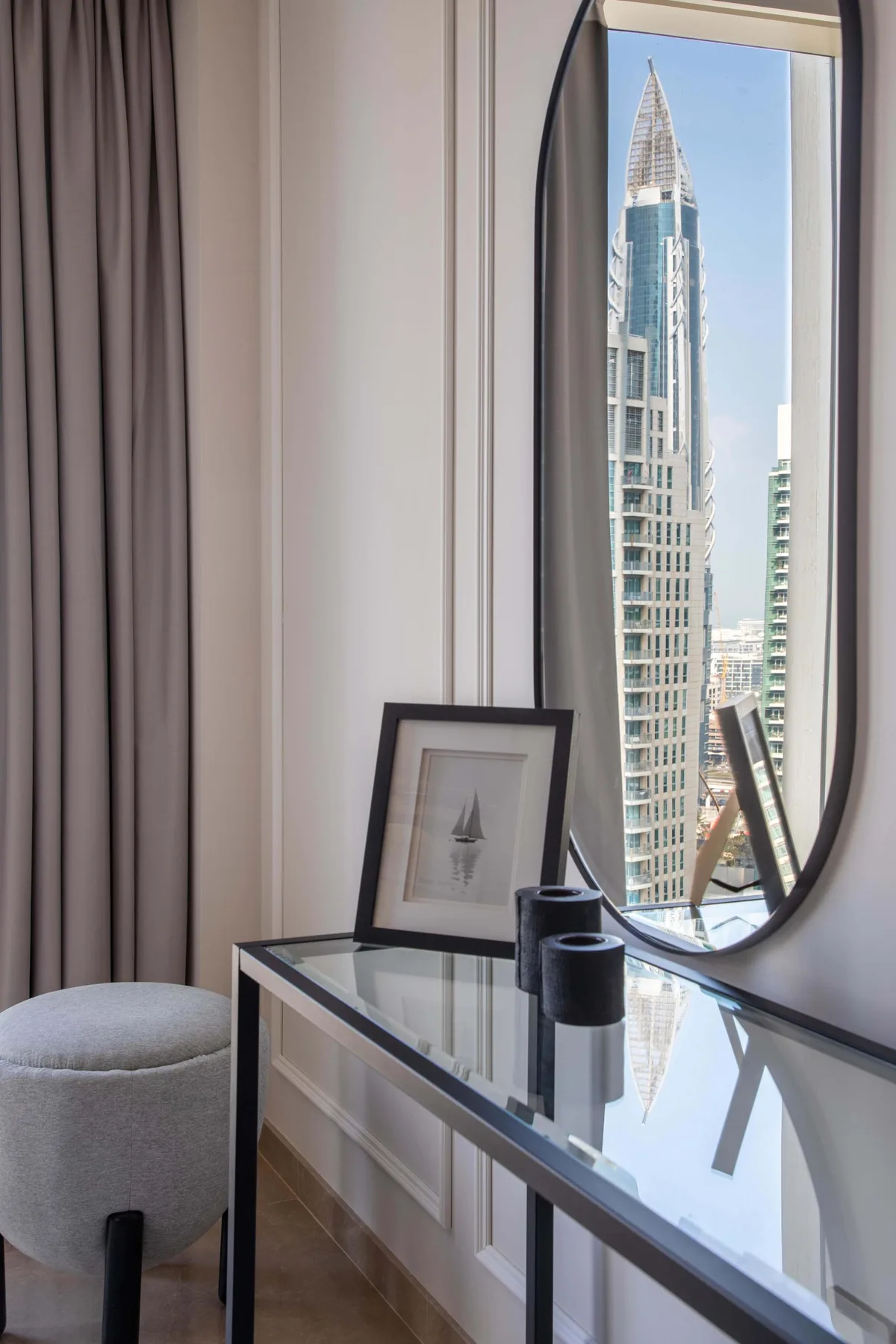 Una habitación luminosa con una consola de vidrio junto a una ventana alta con vista a edificios.