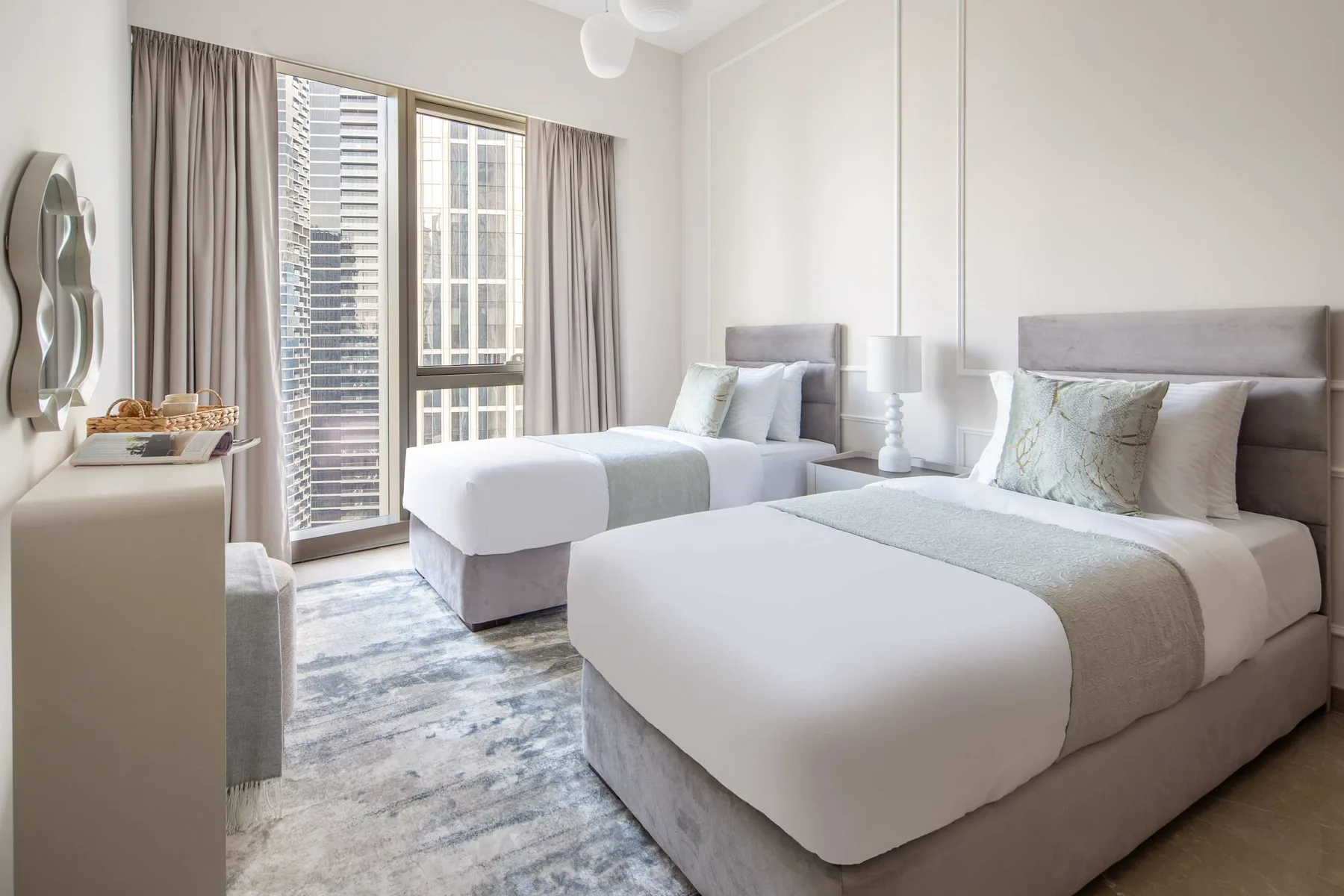 Un dormitorio luminoso con dos camas, ropa de cama clara y ventanas grandes.