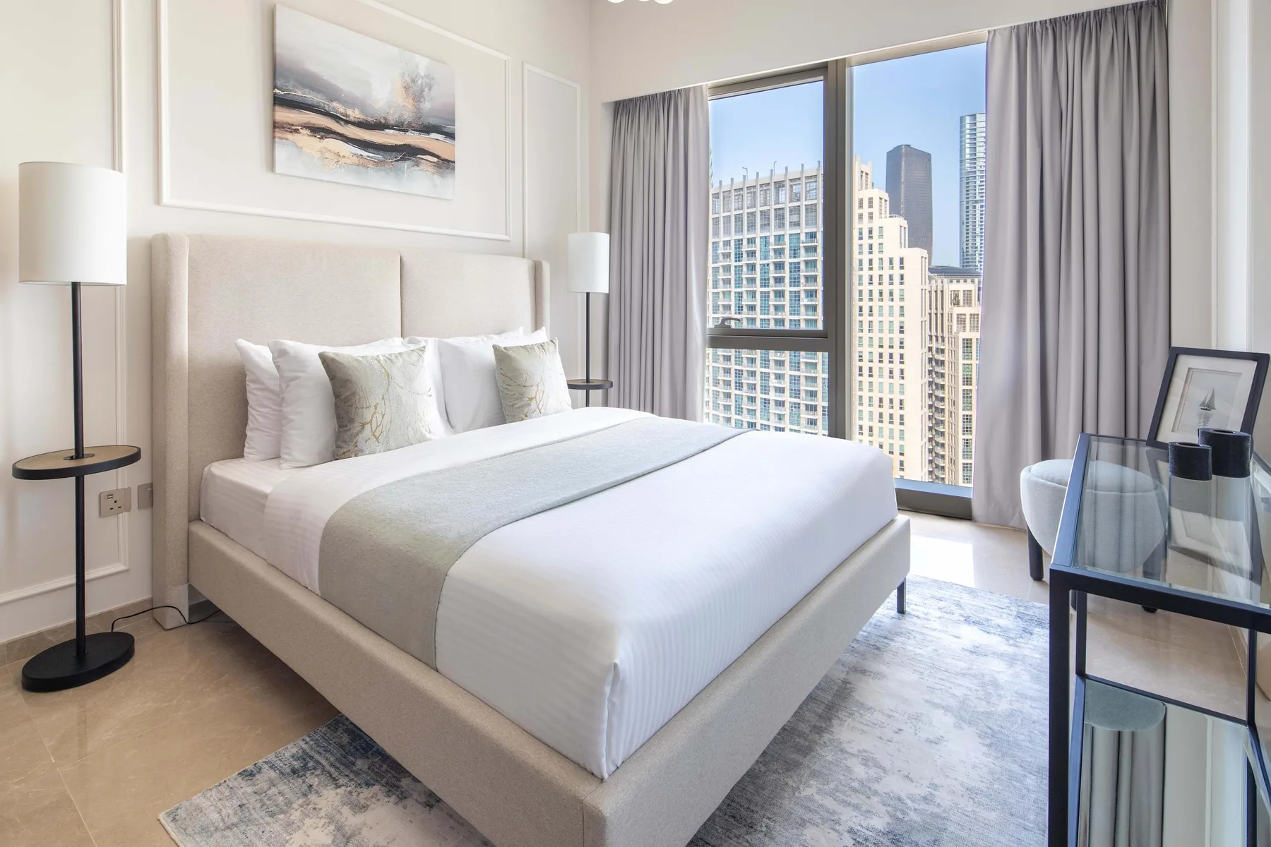 Un dormitorio luminoso con una cama tendida junto a grandes ventanas con vistas a edificios de la ciudad.