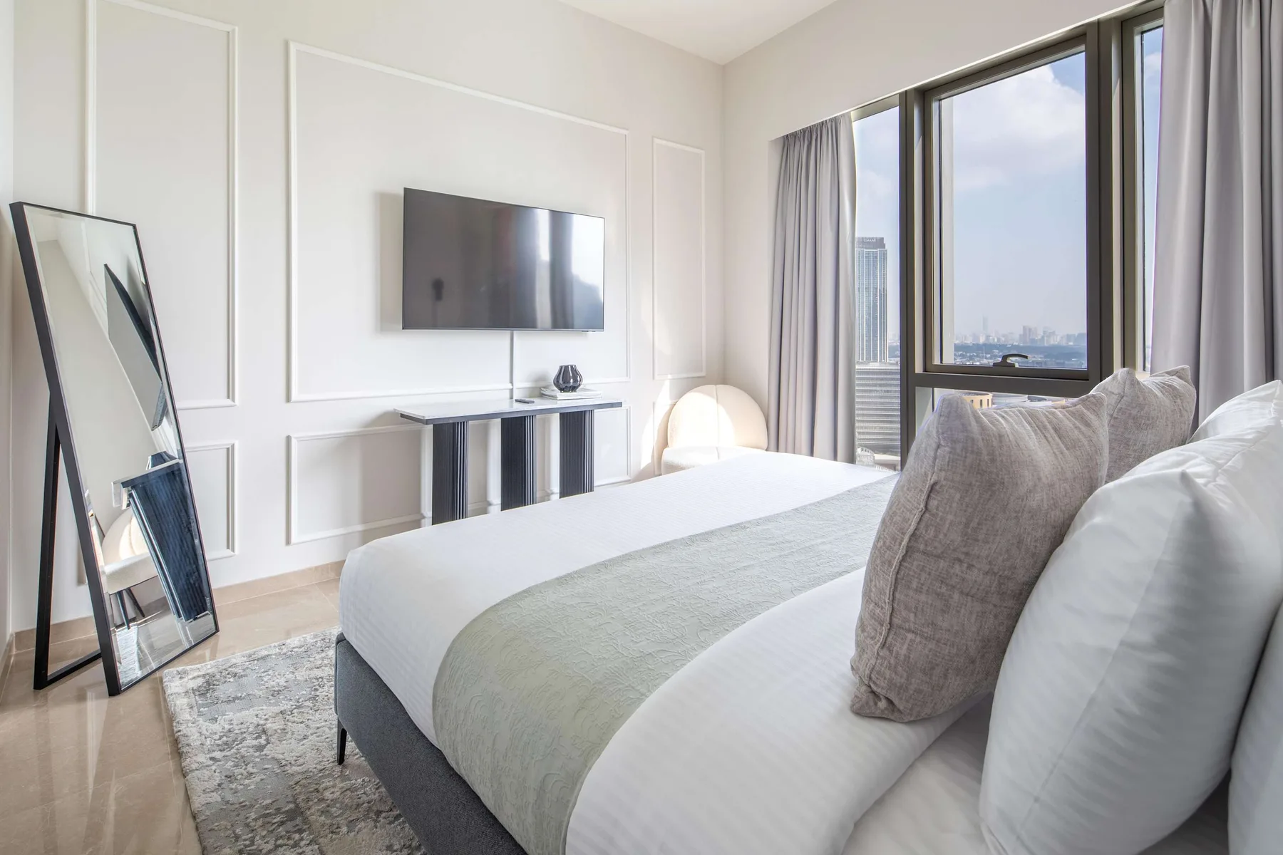 Un dormitorio luminoso con una cama bien tendida, un televisor en la pared y ventanales.