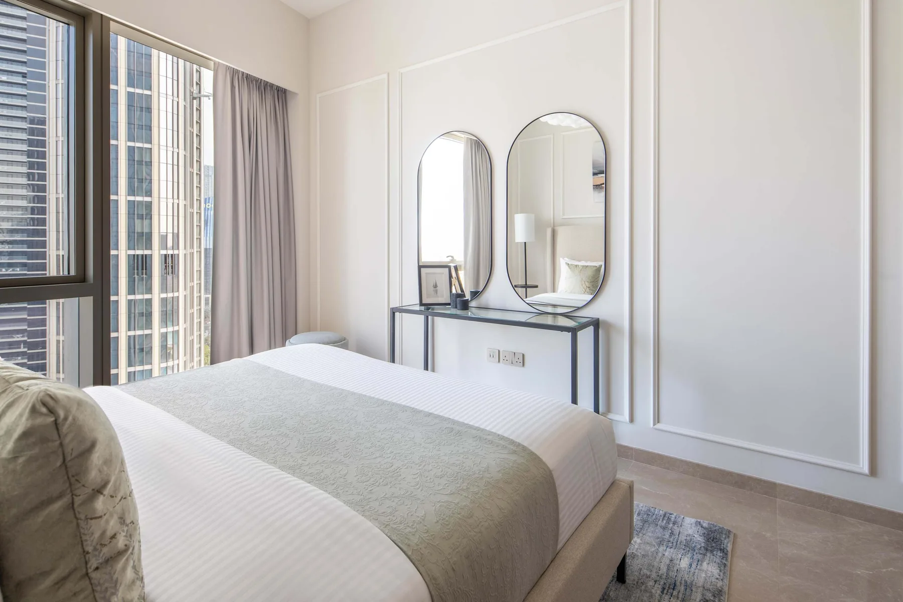 Un dormitorio con cama, tocador con espejos y grandes ventanales.