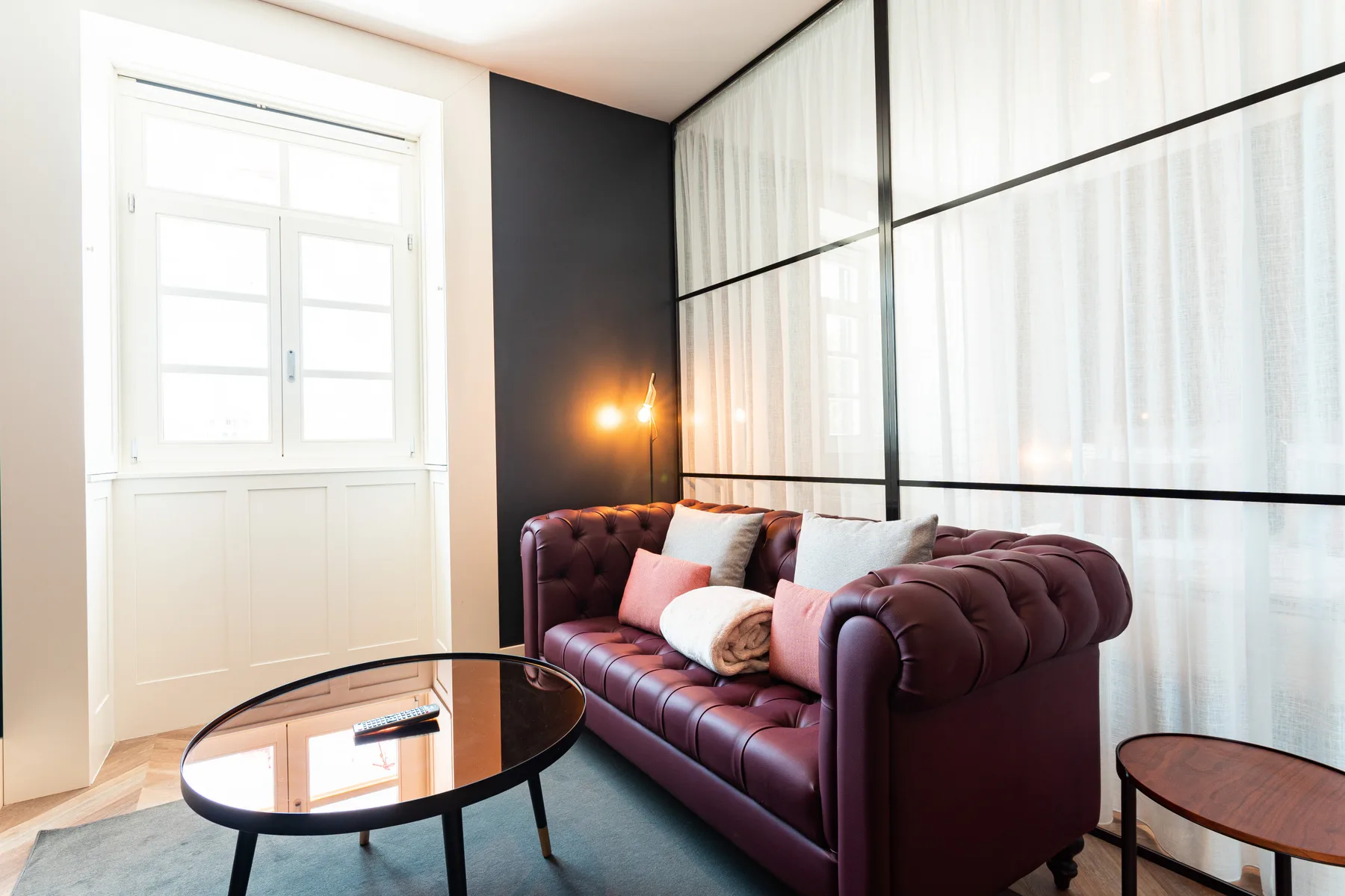 A living room with a burgundy sofa, glass coffee table, and tall curtained windows.