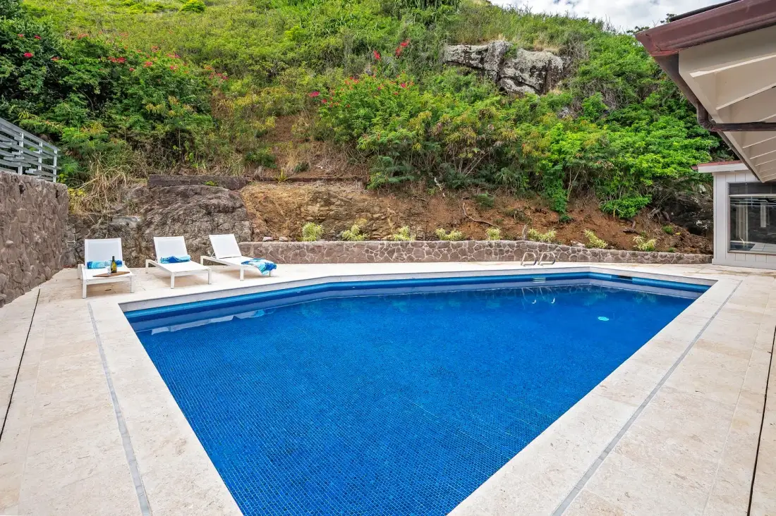 An outdoor swimming pool is surrounded by a paved deck and green hillside.