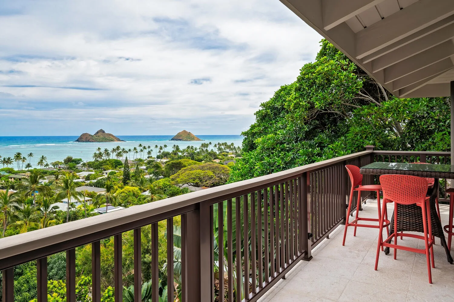 A balcony overlooks a tropical coastline, with outdoor seating beside the railing.