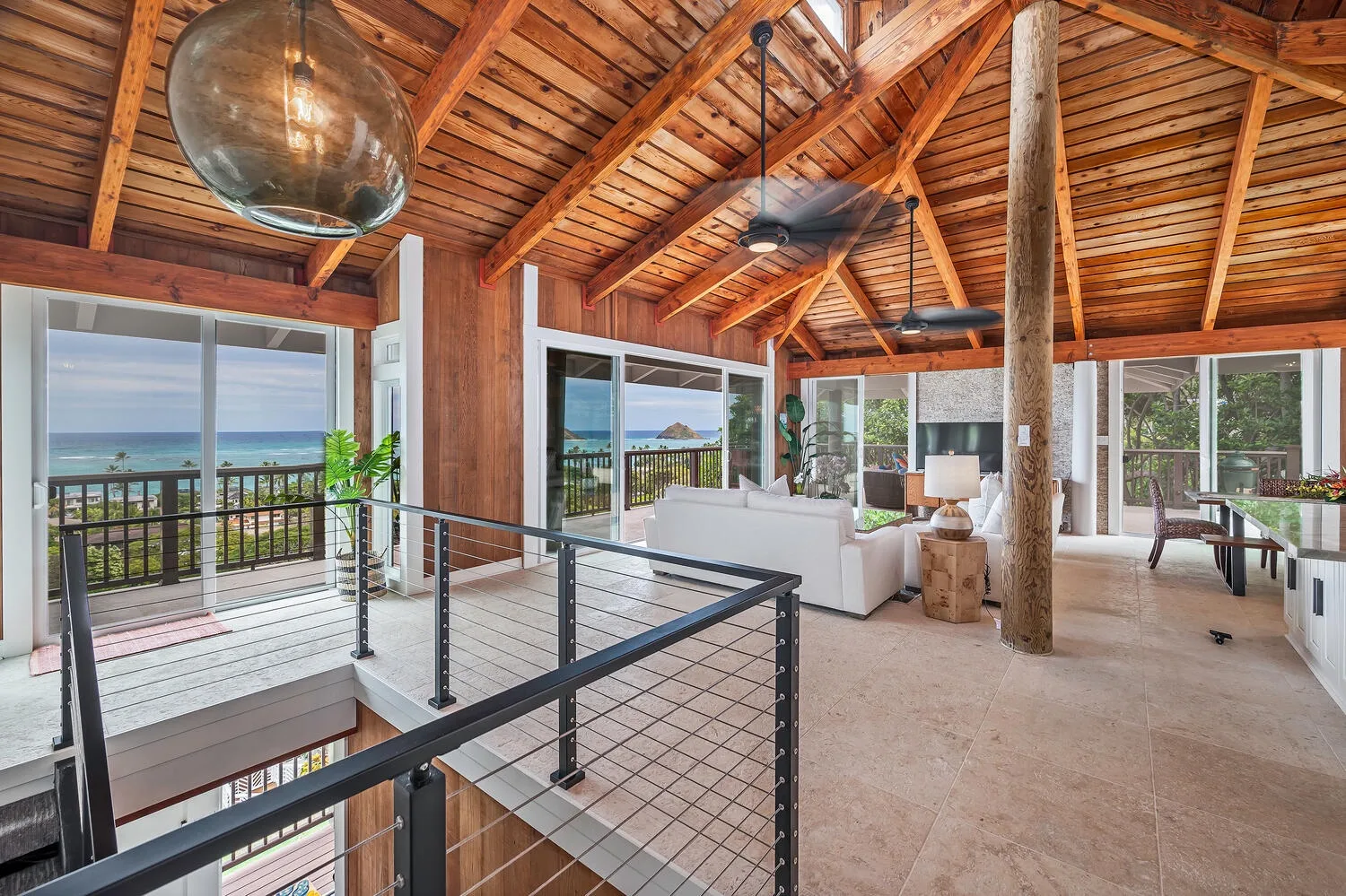 An open living area has a high wooden ceiling, seating, and large windows overlooking the water.