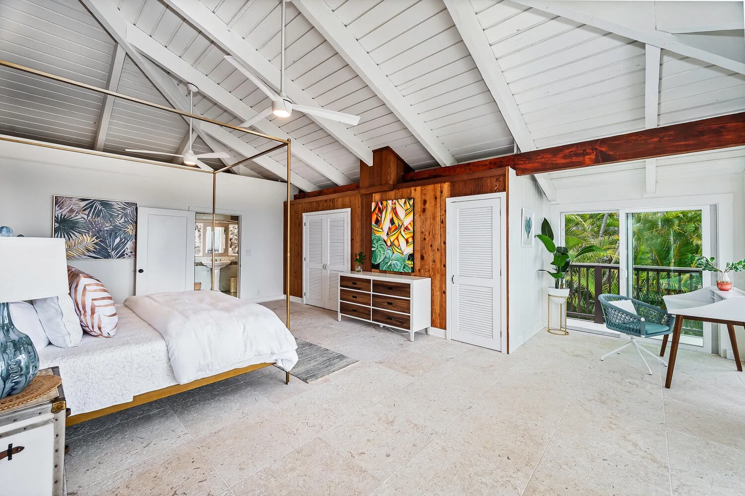 A bright bedroom with a bed, wooden wall panels, and a seating area by the windows.