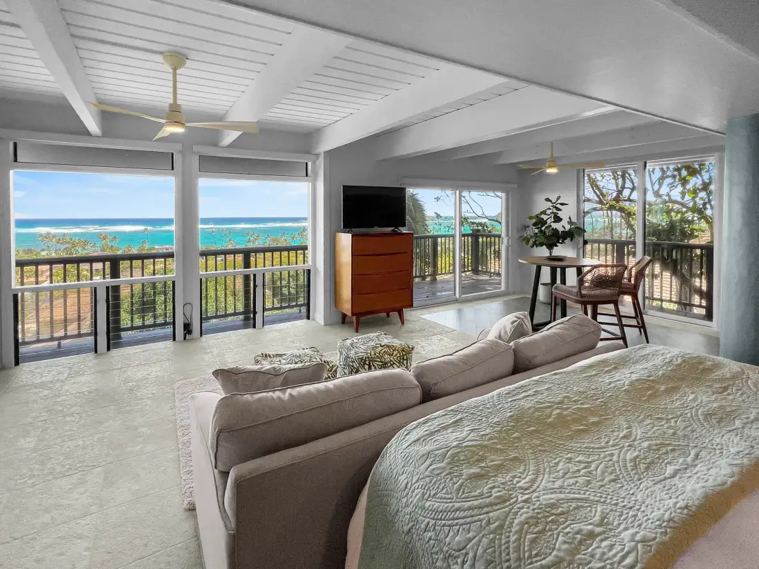 A bright room with a bed, seating, and wide windows overlooking the ocean.