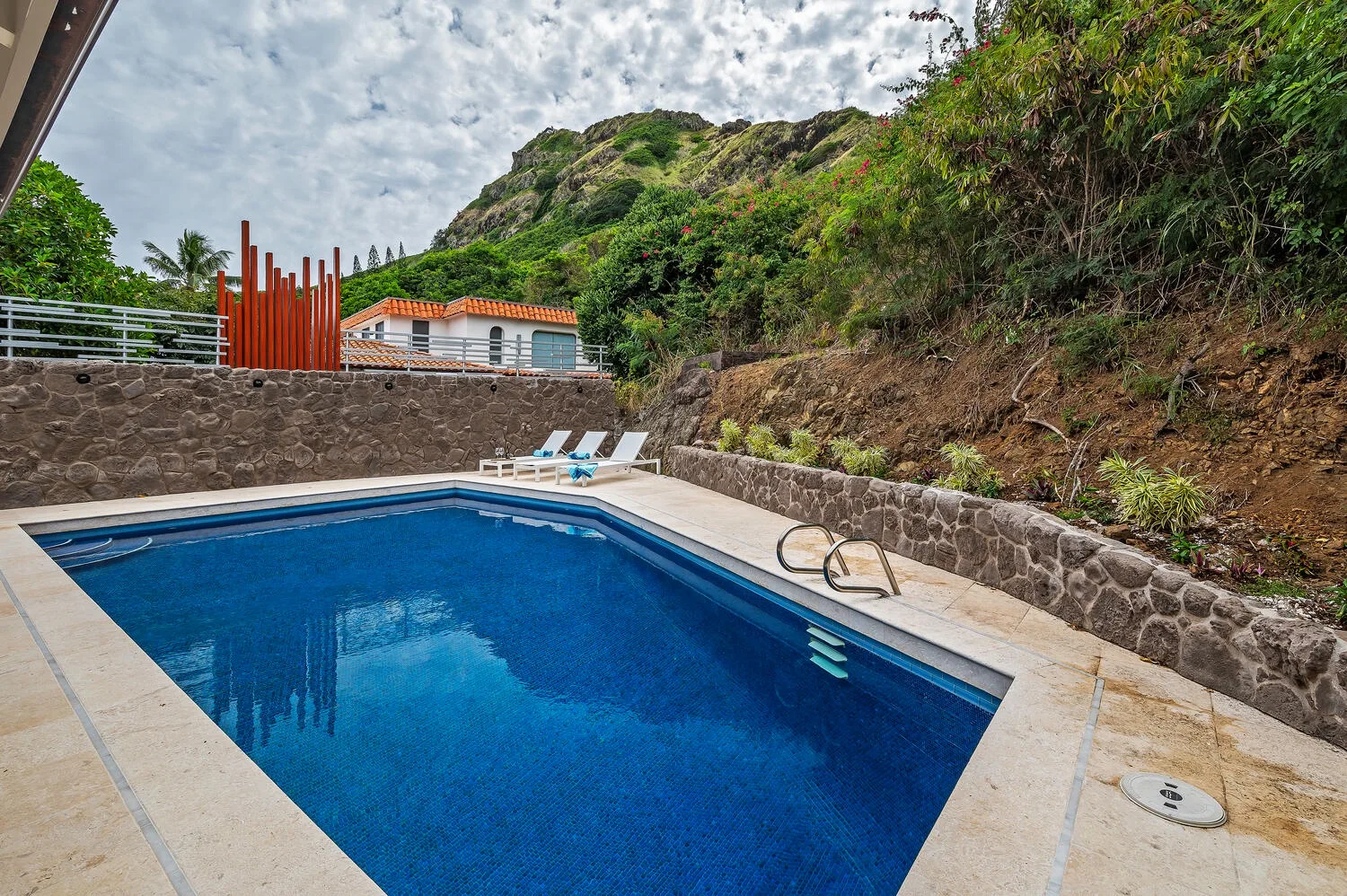 An outdoor swimming pool sits beside a stone wall and green hillside.