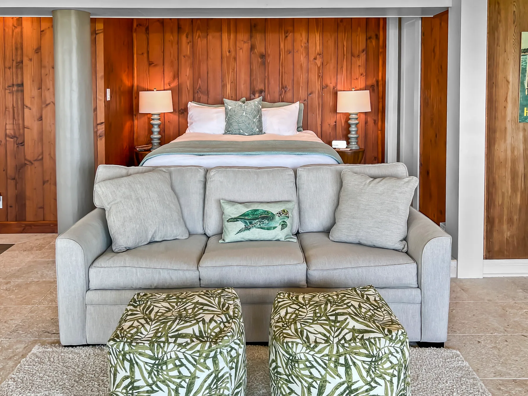 A bedroom with a bed behind a gray sofa and two patterned ottomans.