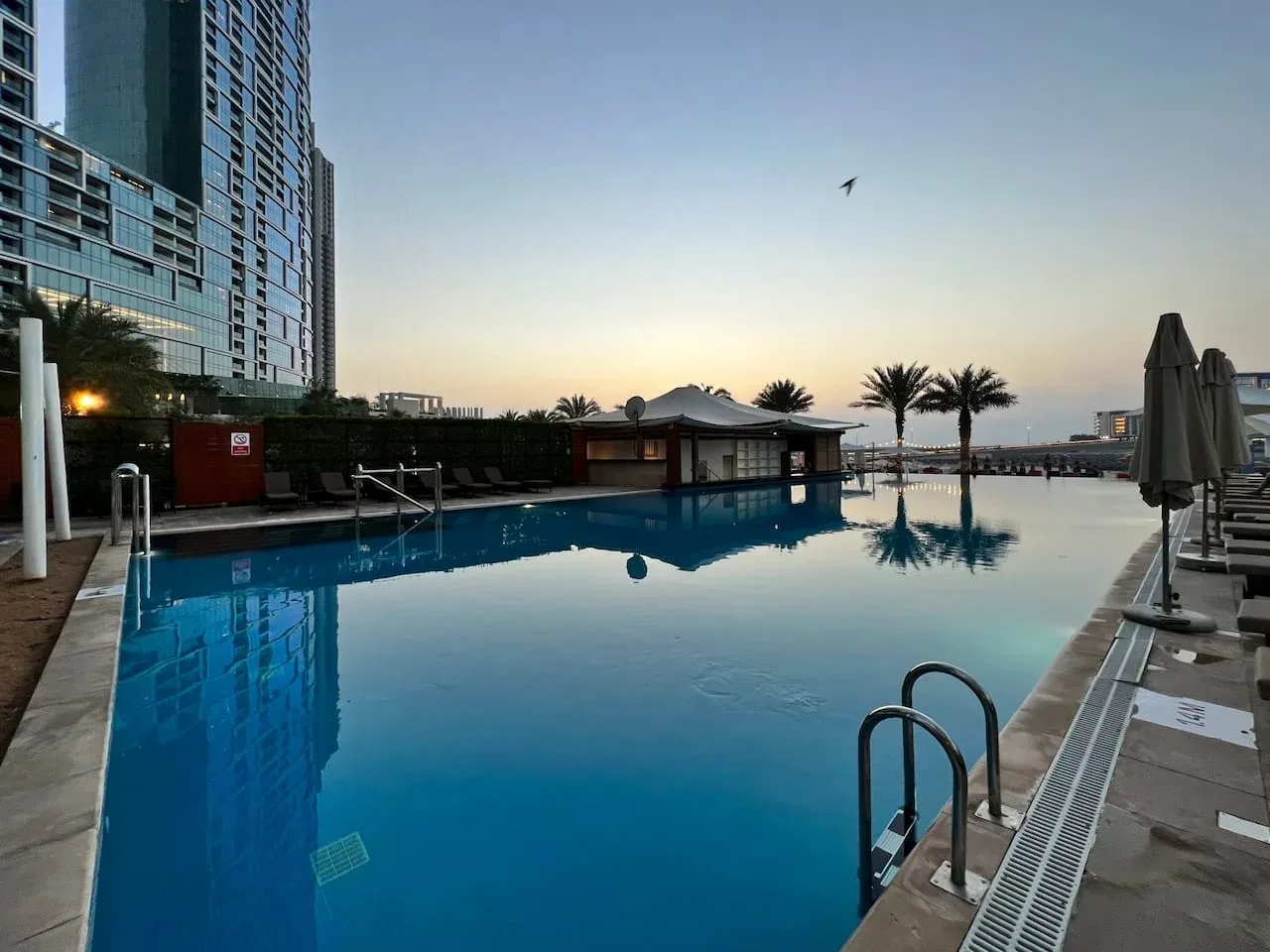 Una piscina al aire libre refleja el cielo del atardecer junto a palmeras y edificios modernos.